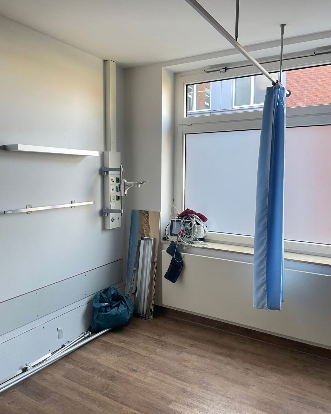 Leeres Patientenzimmer mit Versorgungsleiste, blauem Trennvorhang und abgebauten Geräten auf der Fensterbank